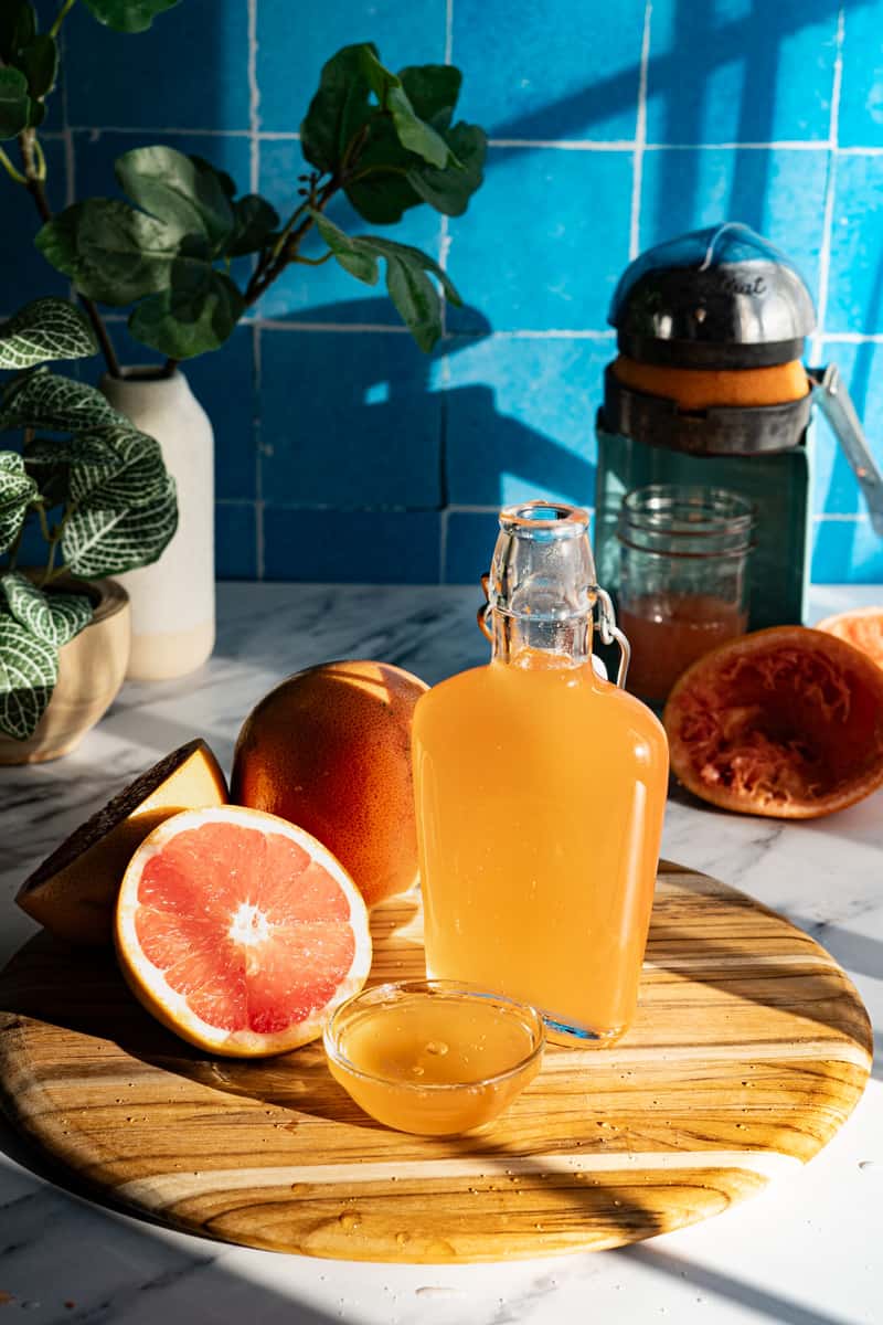 A bottle of homemade grapefruit simple syrup sits on a wooden cutting board on a marble countertop. There is a small dish of simple syrup in front of the bottle and a cut grapefruit to the side of the bottle.