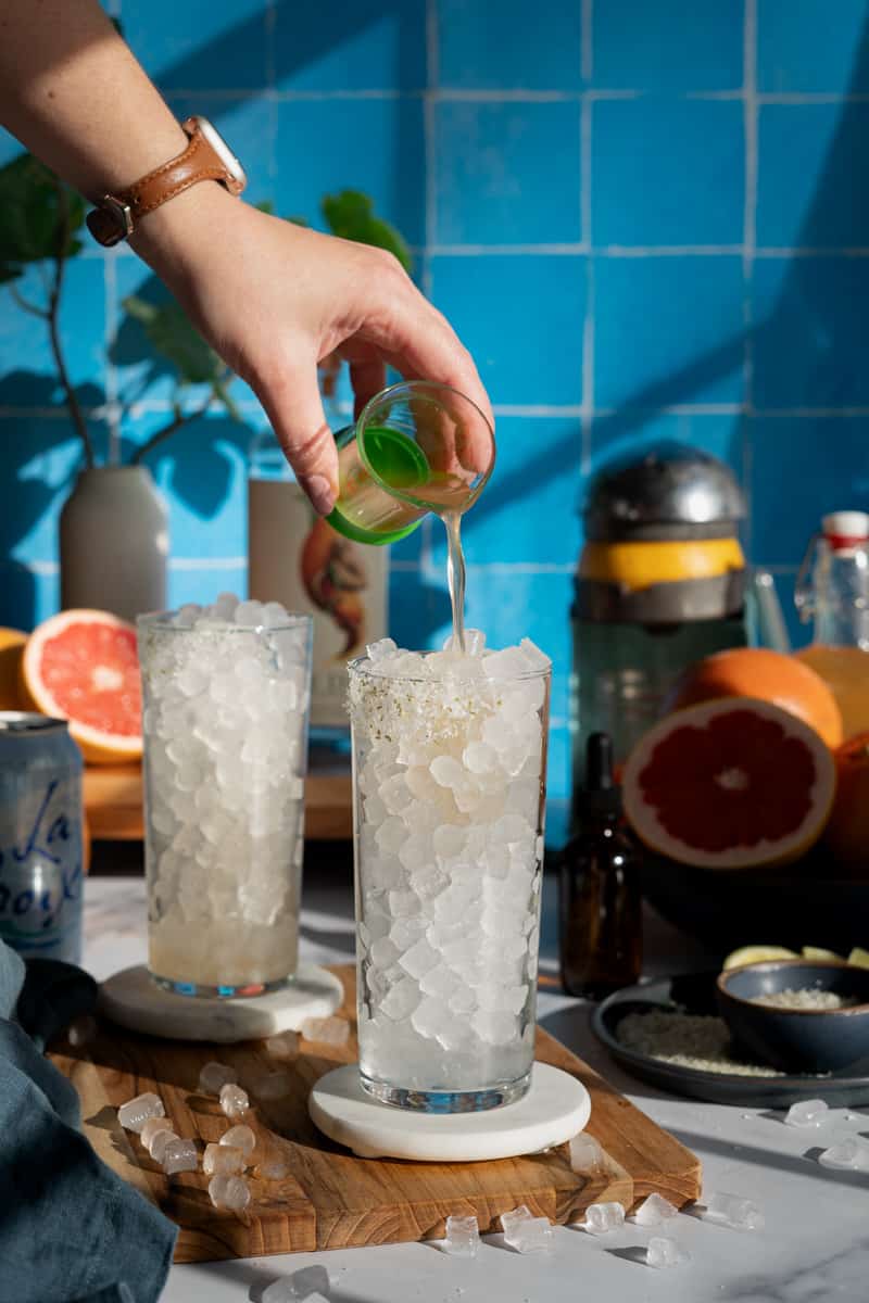 A hand from out of frame is pouring grapefruit simple syrup into a collins glass.