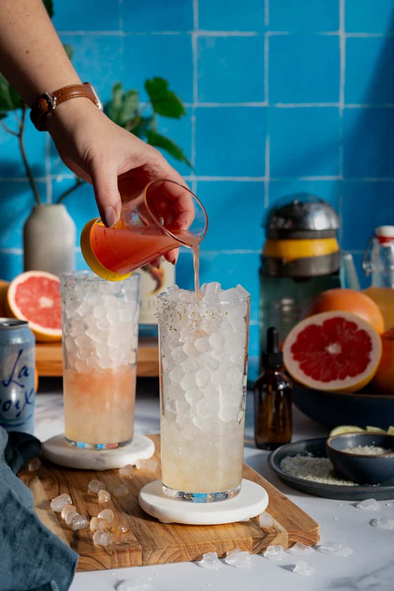Adding grapefruit juice to a glass filled with grapefruit simple syrup to make a paloma mocktail.