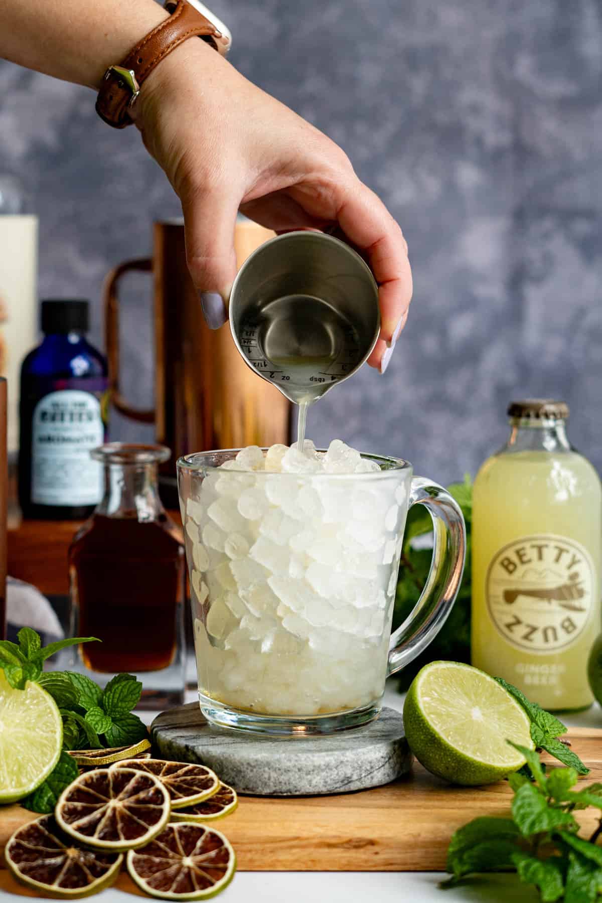 A hand from out of frame is pouring lime juice into a glass mug filled with ice.