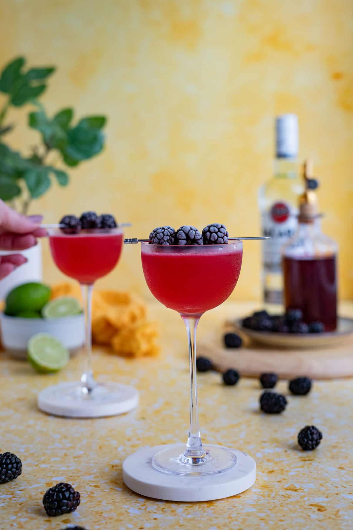 Two blackberry daiquiri cocktails sit on a yellow terrazzo countertop on coasters ready to be served. A hand from out of frame is garnishing one daiquiri with a cocktail pick of frozen blackberries.