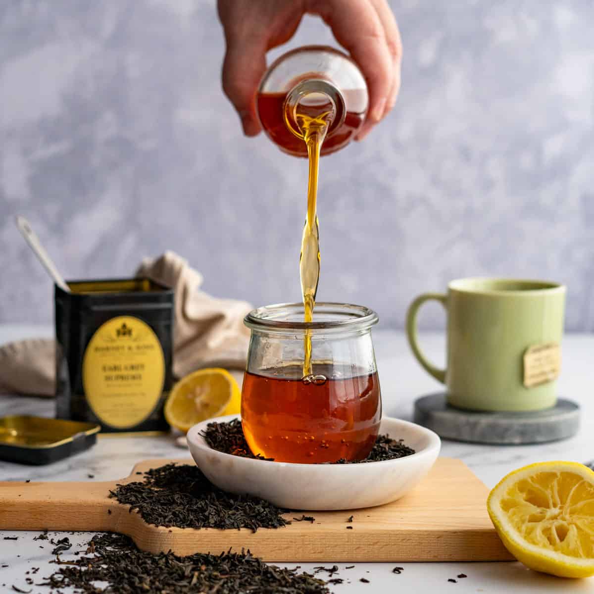 A hand from out of frame is pouring Earl Grey simple syrup into a small glass jar. There is a container of loose leaf Earl Grey tea in the background and a small mug of tea in the background.