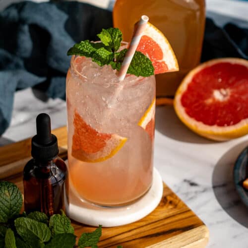A homemade grapefruit Italian soda sits on a wooden cutting board in the sunlight, with a bottle of homemade saline solution to the left of the glass.