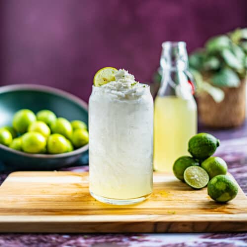 A homemade key lime soda sits on a wooden cutting board with a small pile of key limes next to the glass, and a bottle of key lime simple syrup in the background.