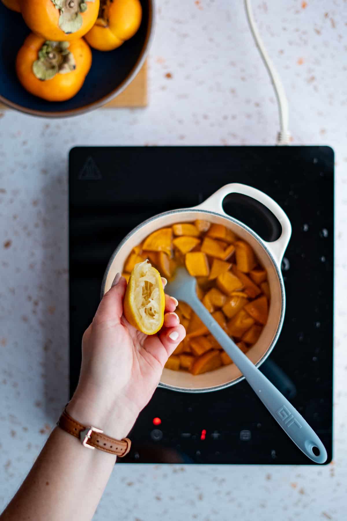 A hand from out of frame is squeezing a lemon into a saucepan of simmering persimmon simple syrup.