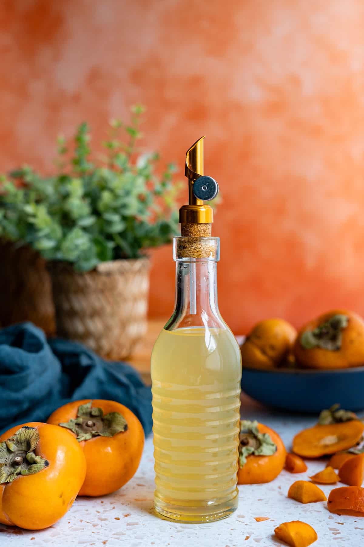 A bottle of persimmon simple syrup sits on a countertop. There are whole and cut persimmons in the frame of the photo as well.