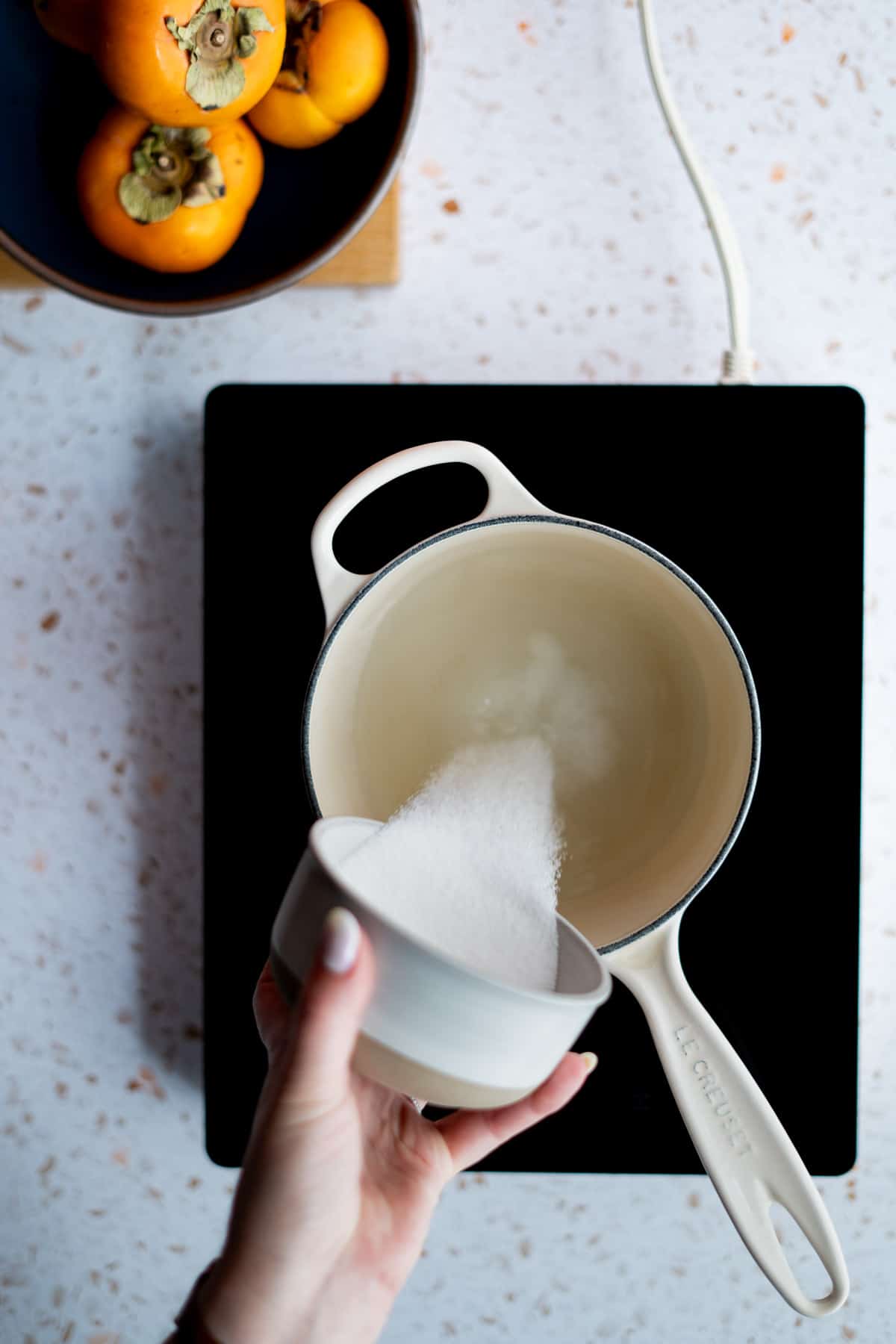A hand from out of frame is pouring a bowl of sugar into a saucepan filled with water.