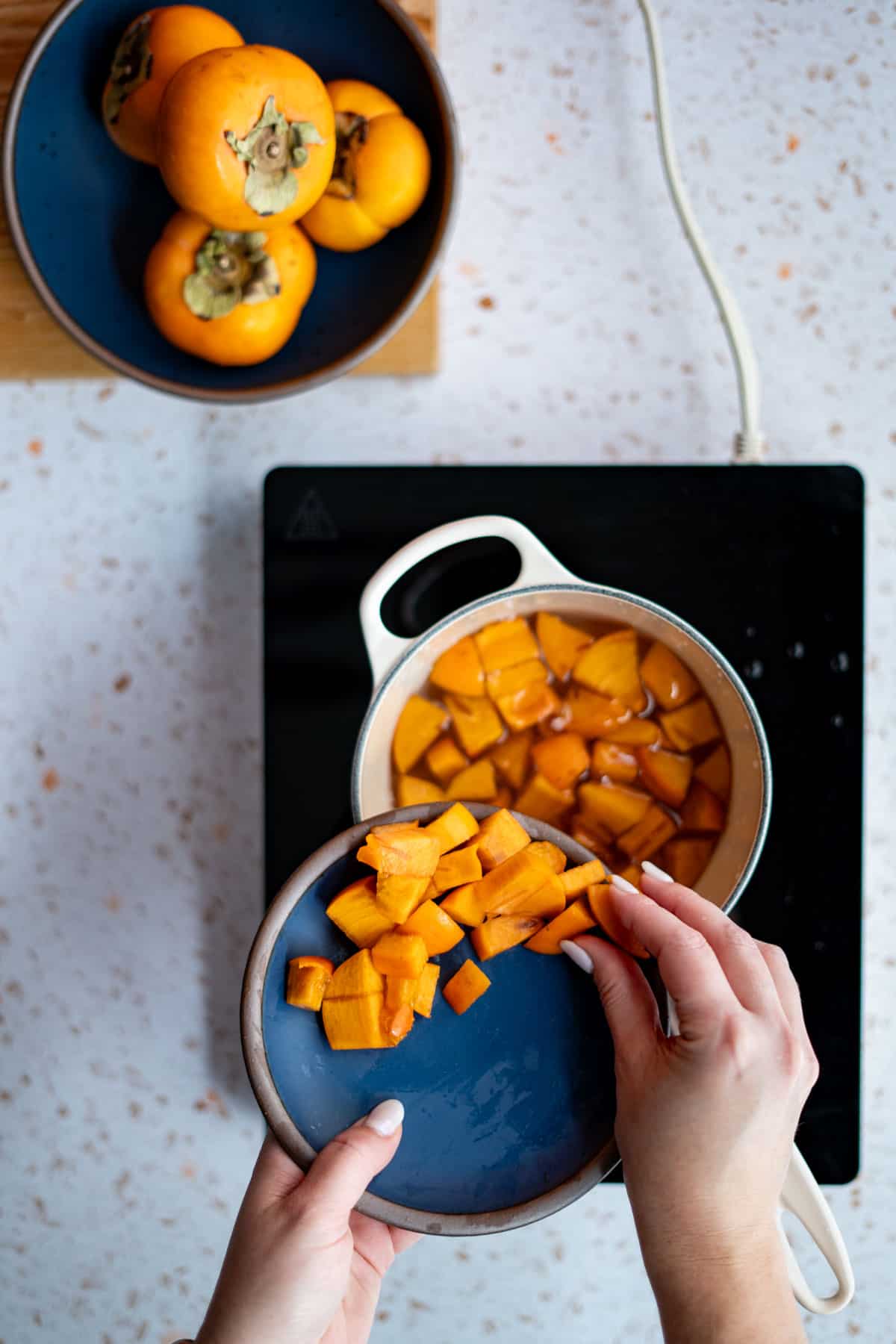 Hands from out of frame are putting cut persimmons into a saucepan of simple syrup.