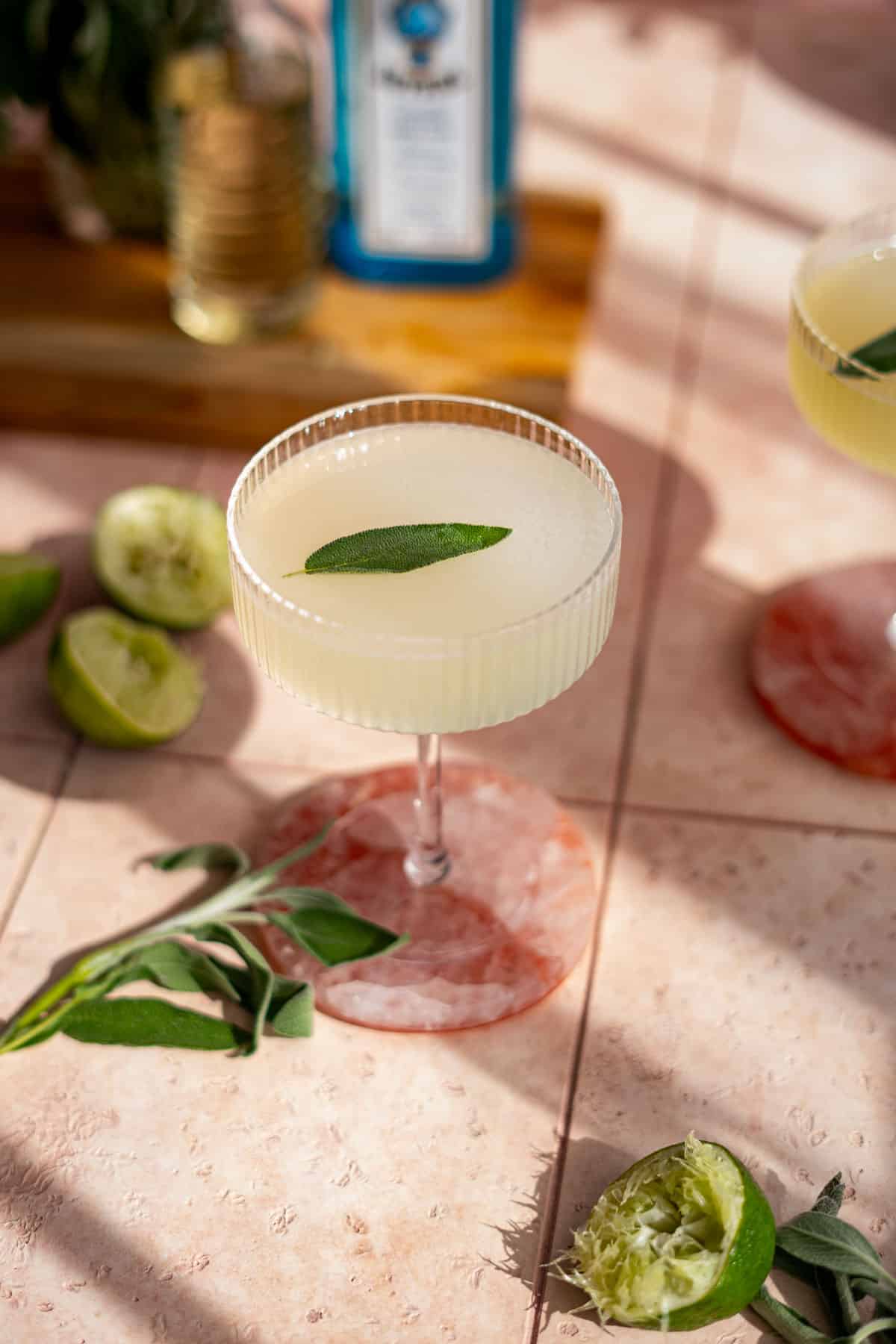 A sage gin gimlet sits on a pink stone countertop with a sprig of sage on the countertop and limes that have been squeezed on the counter.