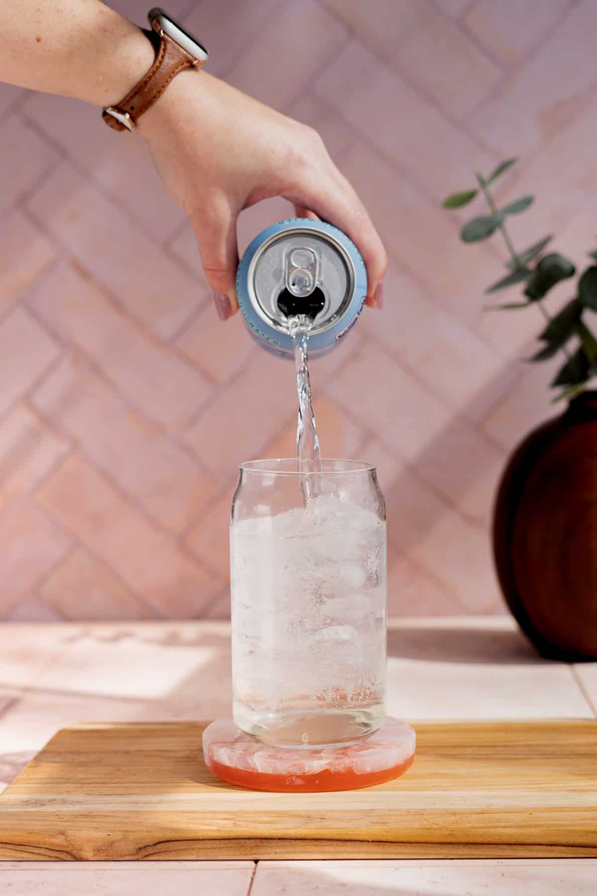 A hand from out of frame is pouring soda water into a soda can glass filled with ice.