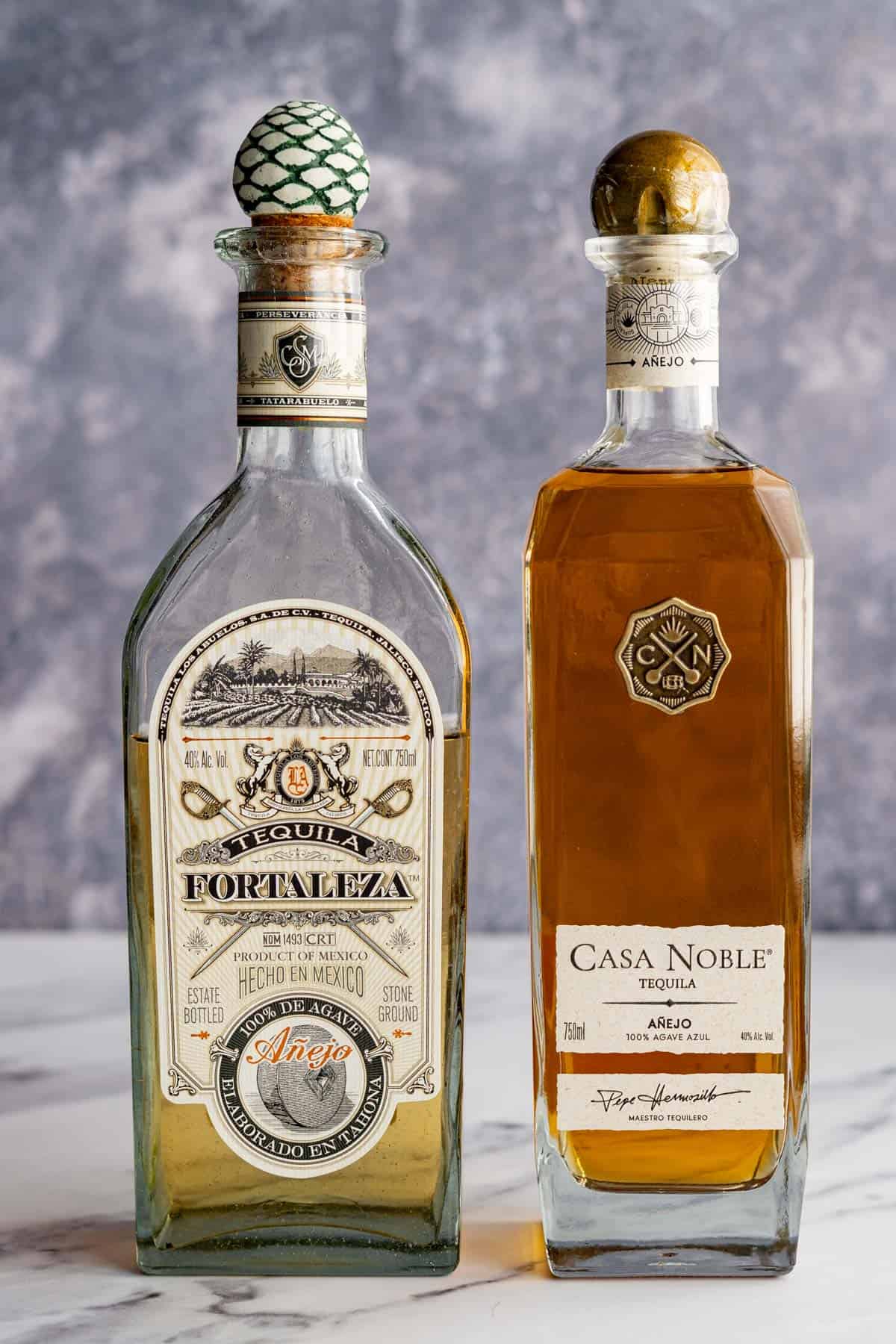 Two bottles of añejo tequila, including Fortuleza and Casa Noble, sit on a white marble countertop.