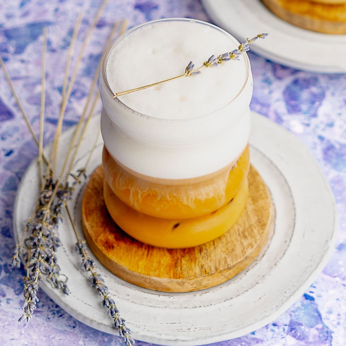 A glass of iced coffee with thick foam on top, garnished with a sprig of lavender, sits on a wooden coaster. Nearby, extra lavender sprigs and a white plate hint at the flavors of a Lavender Honey Latte against a purple textured background.
