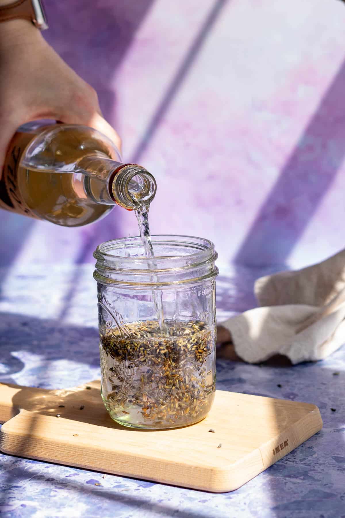 A hand from out of frame is pouring vodka into a jar that has been filled with dried lavender buds.