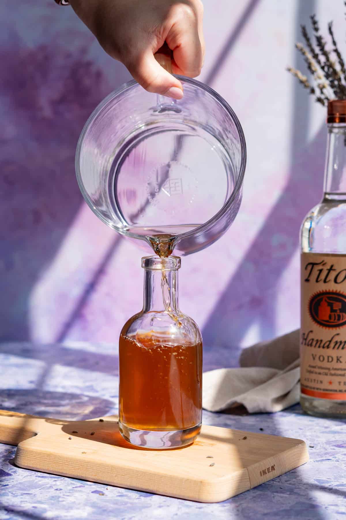 A hand from out of frame is pouring lavender infused vodka into a bottle for storage.