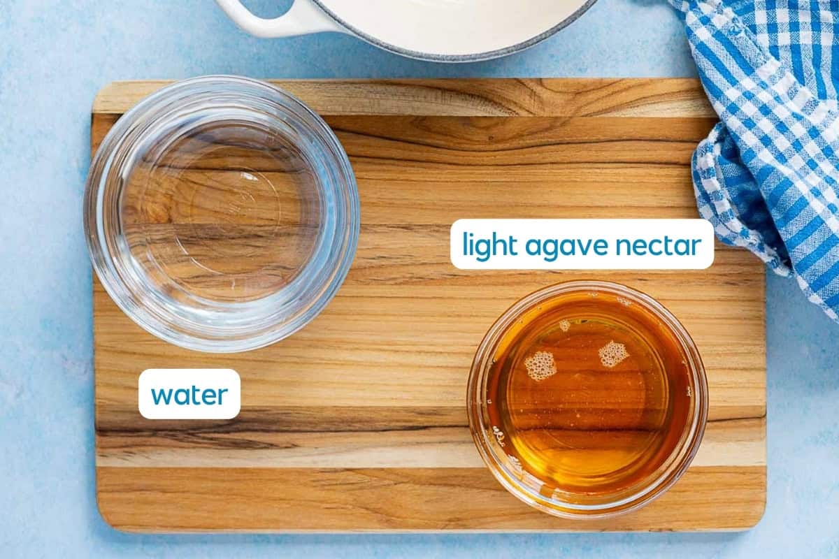 Two glass bowls sit on a wooden board: one with water, the other with light agave nectar, perfect for making agave simple syrup. Each bowl is labeled for clarity. A blue checkered cloth and a white dish are arranged nearby.
