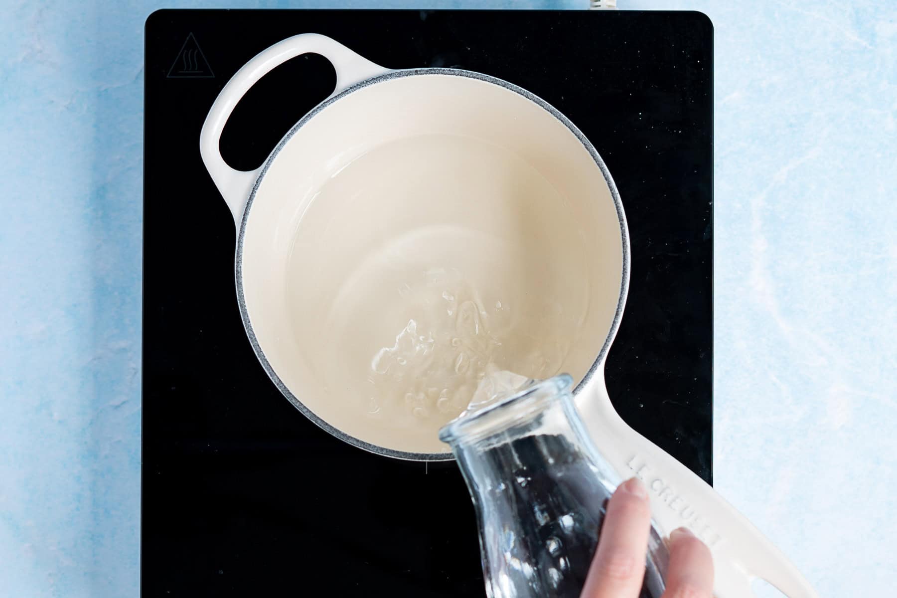 A hand pours water for making agave simple syrup from a glass pitcher into a white pot on a black stovetop, viewed from above.