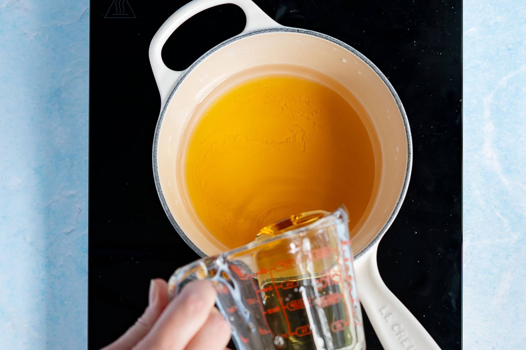 A hand pours agave nectar from a glass measuring cup into a cream-colored pot filled with yellow liquid on a stovetop.