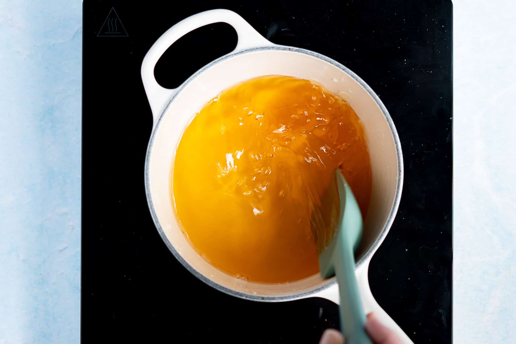 A hand stirs a pot of agave simple syrup on a black stovetop, using a light blue spatula.