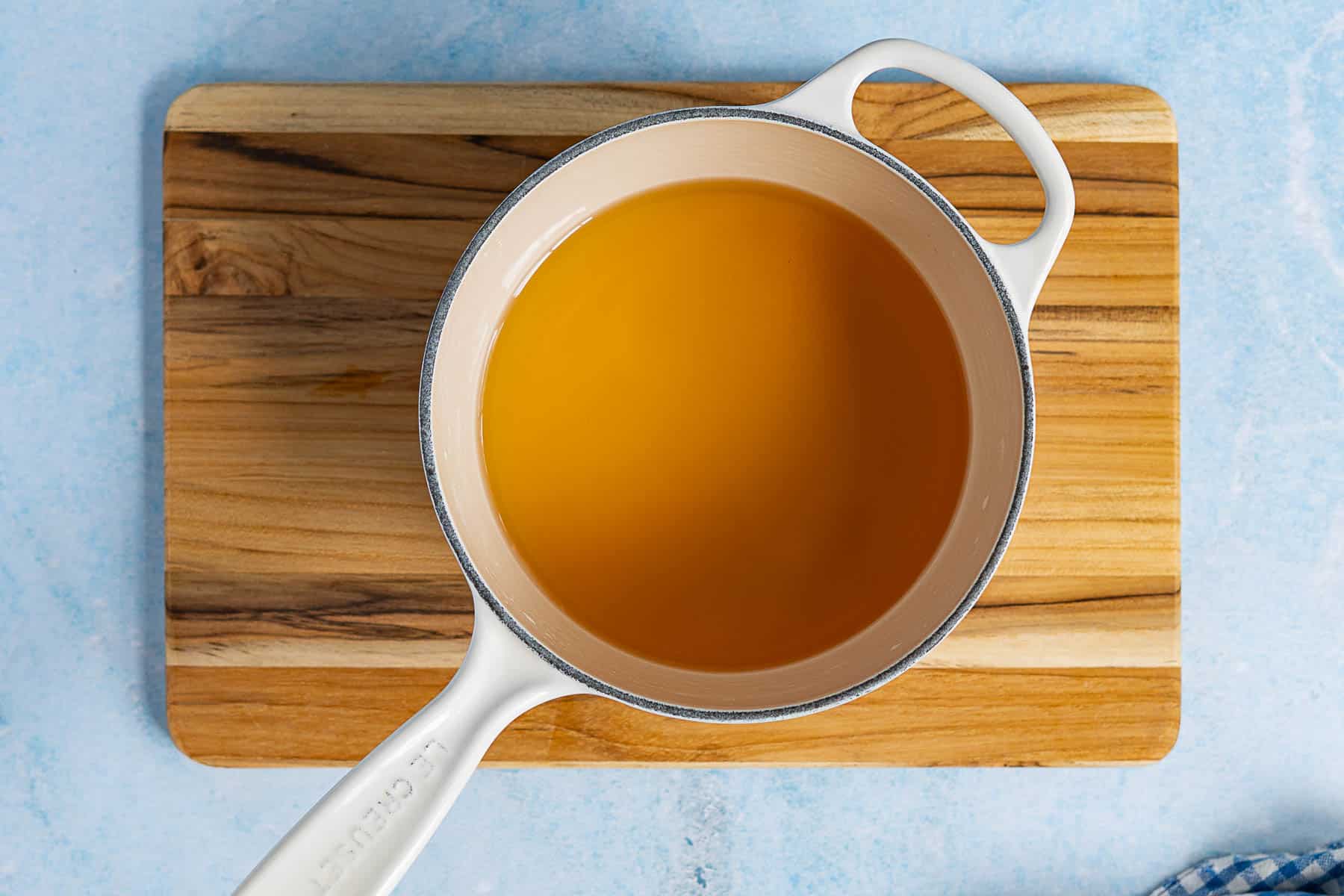 A white saucepan filled with golden agave simple syrup sits on a wooden cutting board against a light blue background.