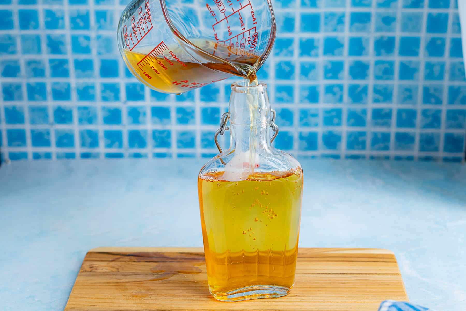 A glass measuring cup pours golden agave simple syrup into a clear glass bottle with a swing top, set on a wooden board against a blue tiled background.