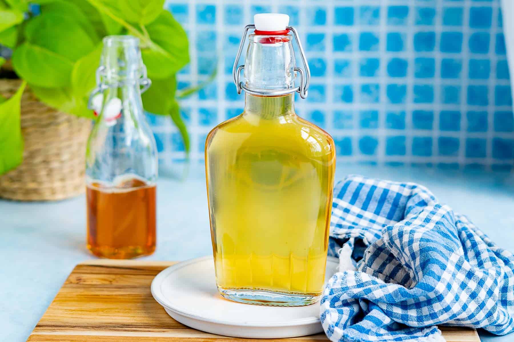 Two glass bottles with metal clasps, one filled with an agave simple syrup, and the other with an agave nectar, sit on a wooden board with a blue checkered cloth beside them and a leafy plant in the background.