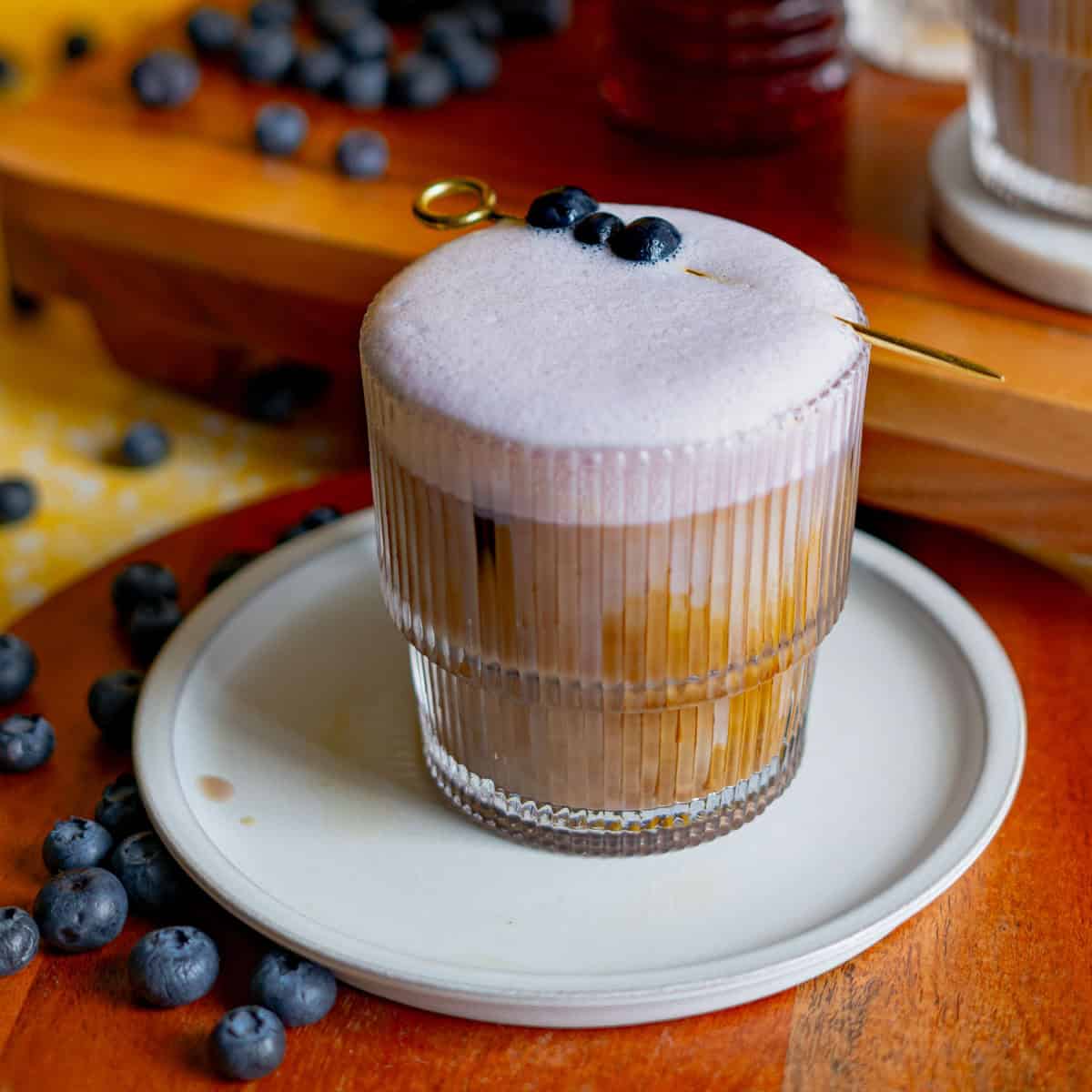 A blueberry latte is garnished with a cocktail pick of blueberries. Blueberries are scattered around the frame.