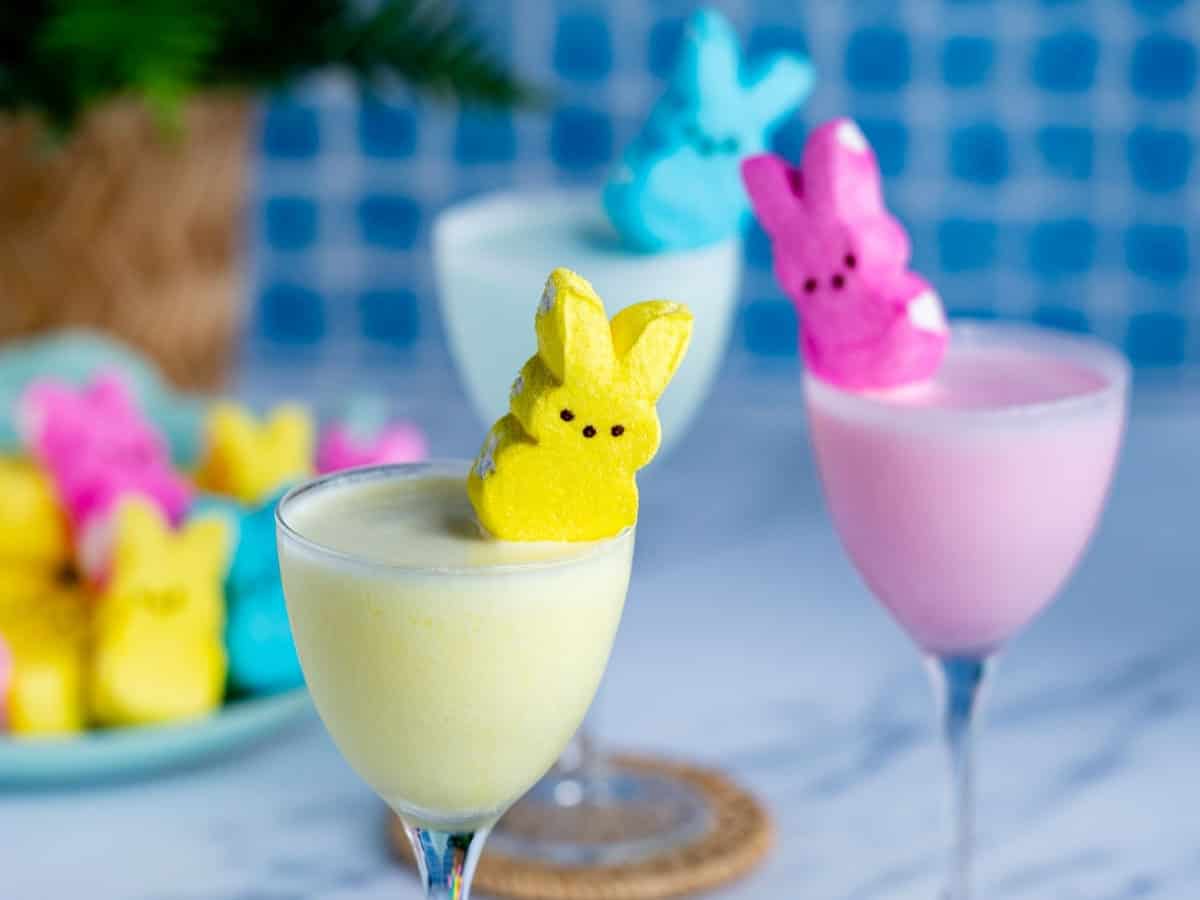 Three pastel-colored peeps martini drinks in cocktail glasses, each topped with a marshmallow bunny Peep. In the background, more Peeps are arranged on a plate. The setting is bright and festive with a blue tiled backdrop.