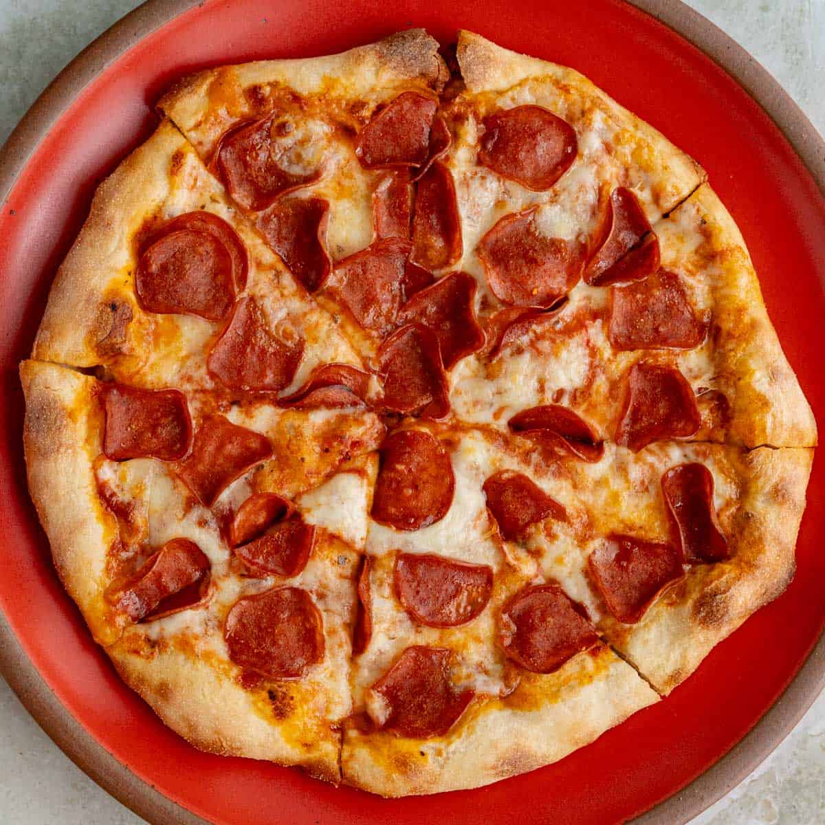 A turkey pepperoni pizza with a golden-brown crust sits on a red plate. Crispy slices of turkey pepperoni are layered on the melted cheese, evenly spread across the pie. Cut into eight slices, it boasts a slightly irregular edge.