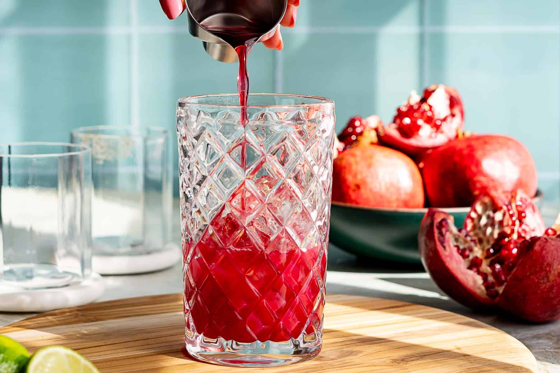 A hand pours pomegranate juice from a small metal cup into a textured glass on a wooden surface. A whole pomegranate and lime appear blurred in the background against a light blue tiled wall, hinting at the freshness of a pomegranate margarita. Sunlight casts shadows across the scene.
