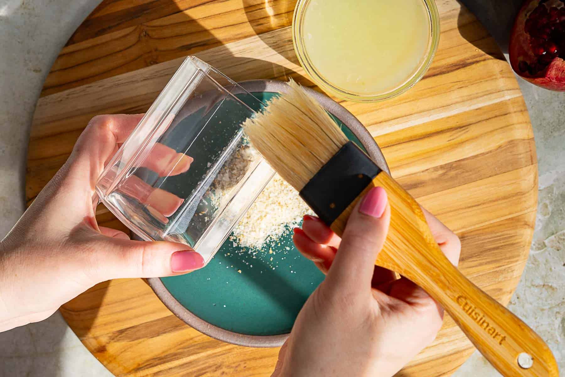 A person uses a wooden culinary brush to sweep breadcrumbs from a container onto a round green plate on a wooden board. Beside it, a small bowl of liquid suggests the presence of something delightful, like the vibrant richness of pomegranate margarita. The scene is well-lit, highlighting textures and details.