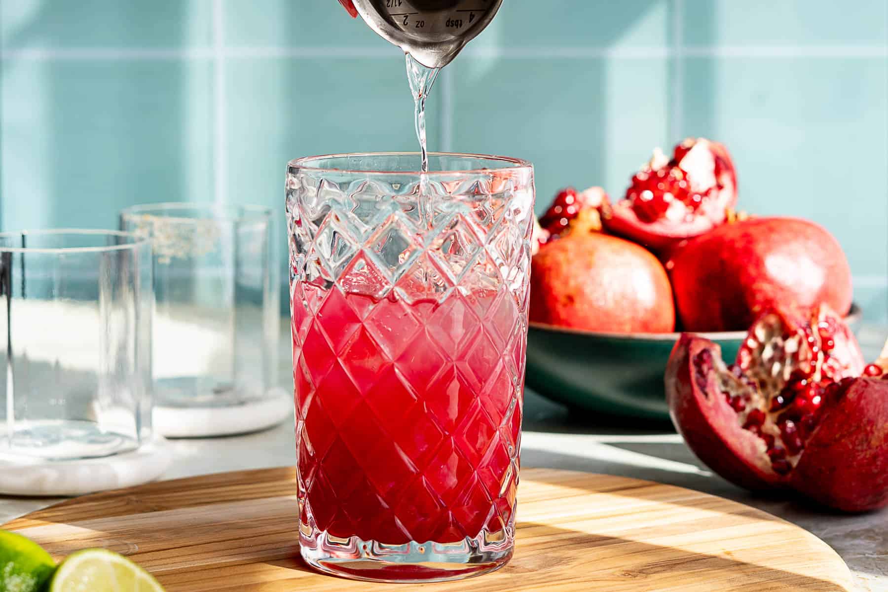 A hand pours silver tequila from a jigger into a textured glass filled with a red pomegranate margarita. The background showcases a bowl of pomegranates against a tiled wall. The brightly lit scene highlights the vibrant ingredients and inviting atmosphere.
