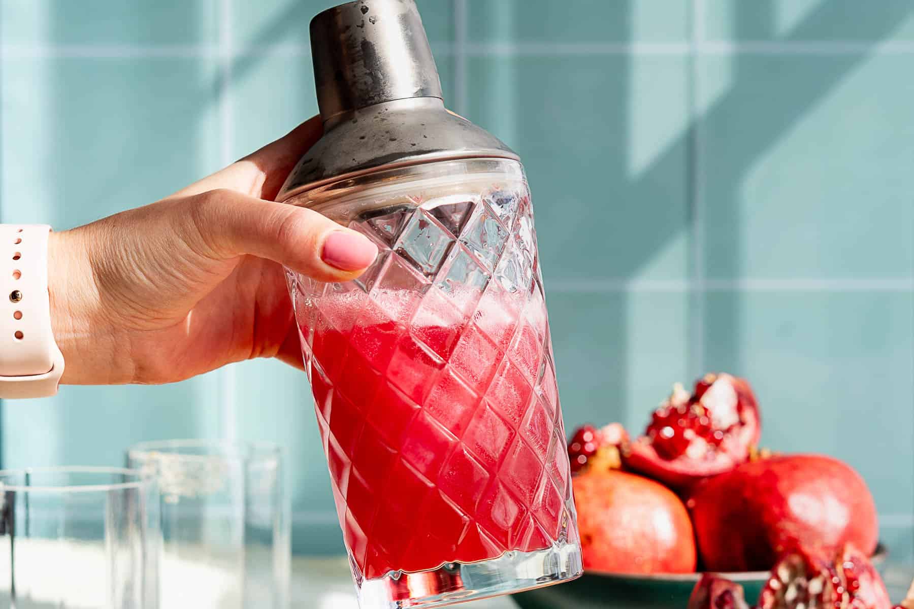 A hand holds a glass cocktail shaker brimming with a vibrant pomegranate margarita. Pomegranates rest in a bowl in the background, with glasses on the counter. The scene is set against a teal tiled wall.