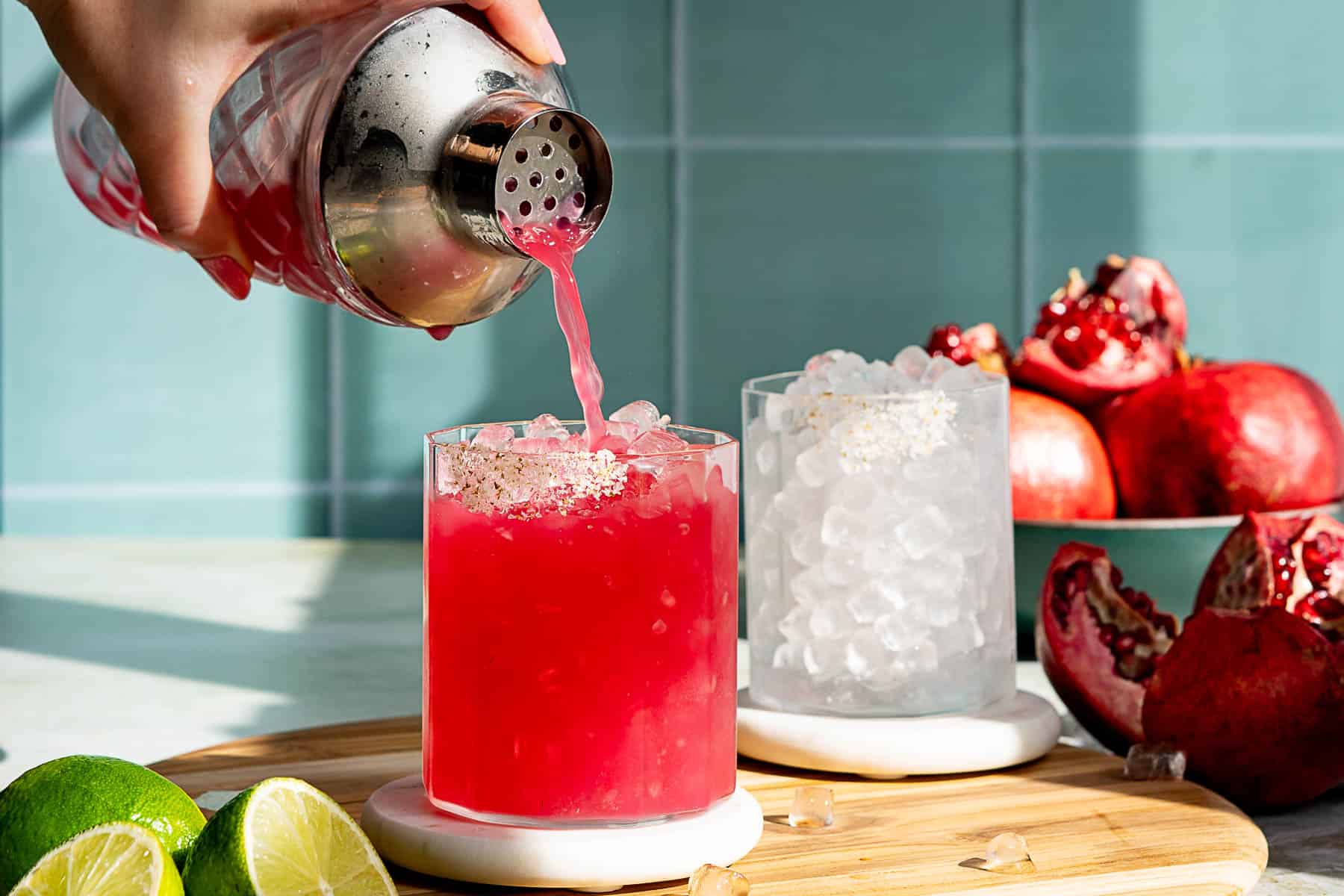 A person pours a pink cocktail from a shaker into a glass filled with ice on a wooden board. Nearby, pomegranate pieces and a lime half suggest it might be a pomegranate margarita. A teal tiled wall serves as the background, and a second glass is in the scene.