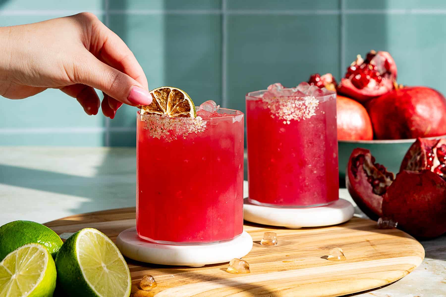 A person garnishes a vibrant red pomegranate margarita with a lime slice in a glass filled with ice. Another similar cocktail is in the background on the wooden board. A halved lime and ice cubes are placed nearby on the table.