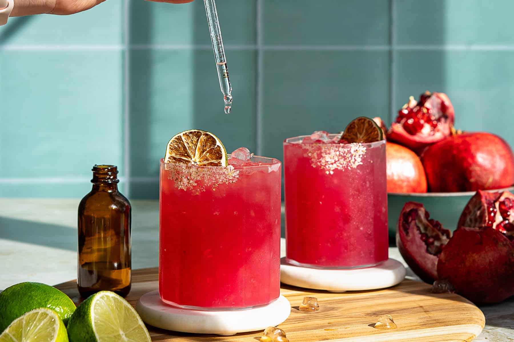 A person garnishes a vibrant red pomegranate margarita with a lime slice in a glass filled with ice. Another similar cocktail is in the background on the wooden board. A halved lime and ice cubes are placed nearby on the table.