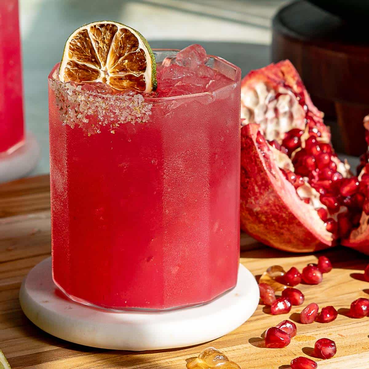 A vibrant pomegranate margarita cocktail in a glass with salted rim, garnished with a dried lime slice. Its placed on a round coaster. Nearby, theres a halved pomegranate and scattered seeds on a wooden background.