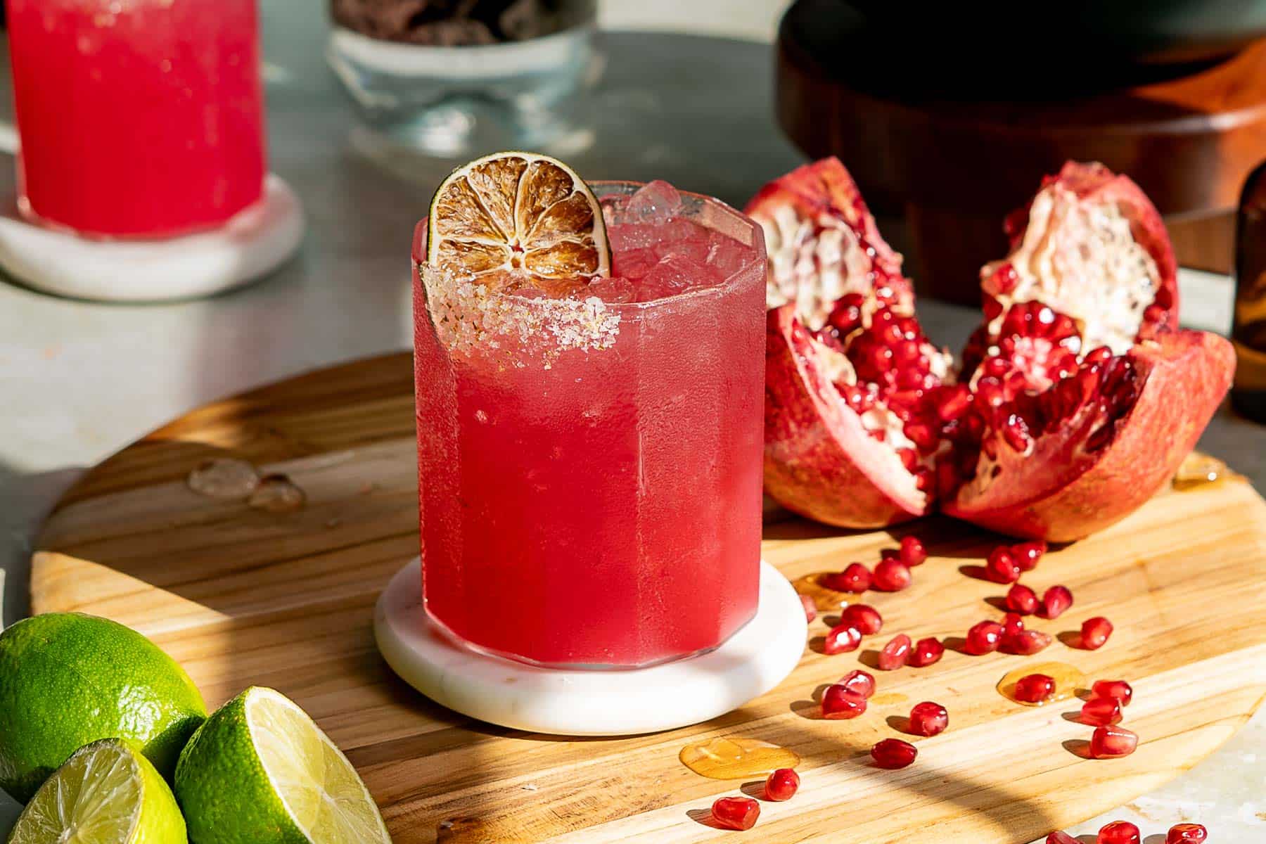 A red pomegranate margarita with ice and a dried lime slice garnish sits on a wooden board, surrounded by pomegranate seeds, a halved pomegranate, and lime wedges. Sunlight casts shadows, creating a fresh and inviting atmosphere.