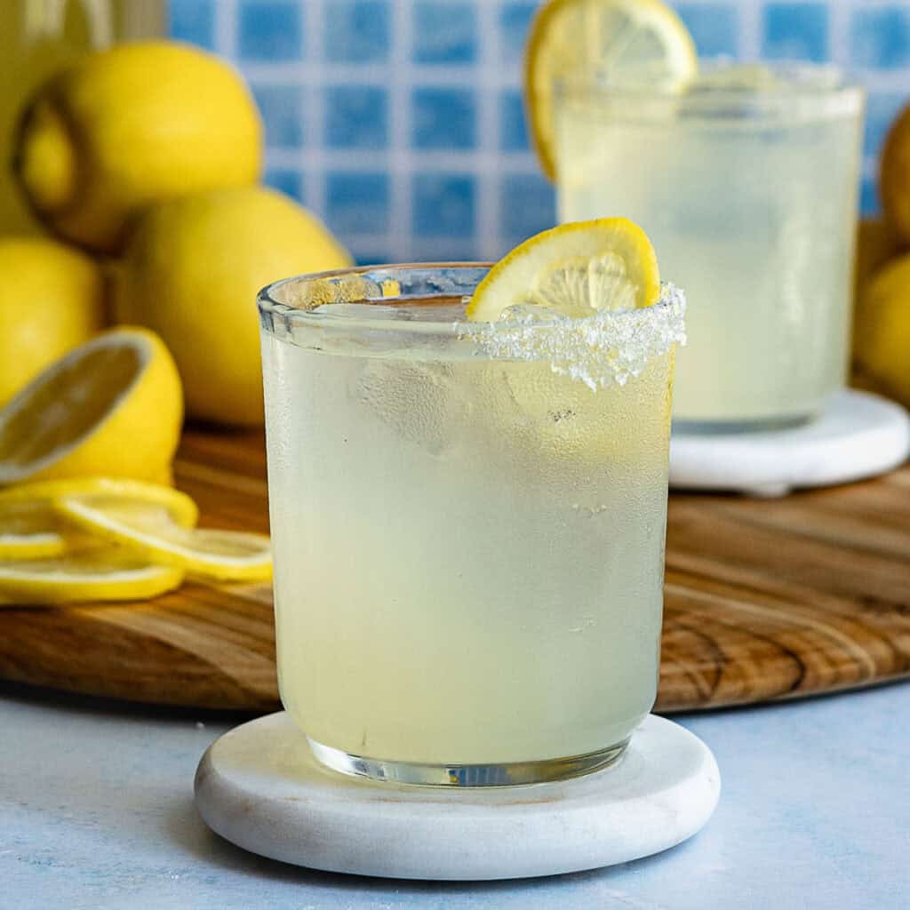 A glass of limoncello margarita with ice and a sugared rim, garnished with a lemon slice, sits on a coaster. Another glass and several whole and sliced lemons are in the background on a wooden board.