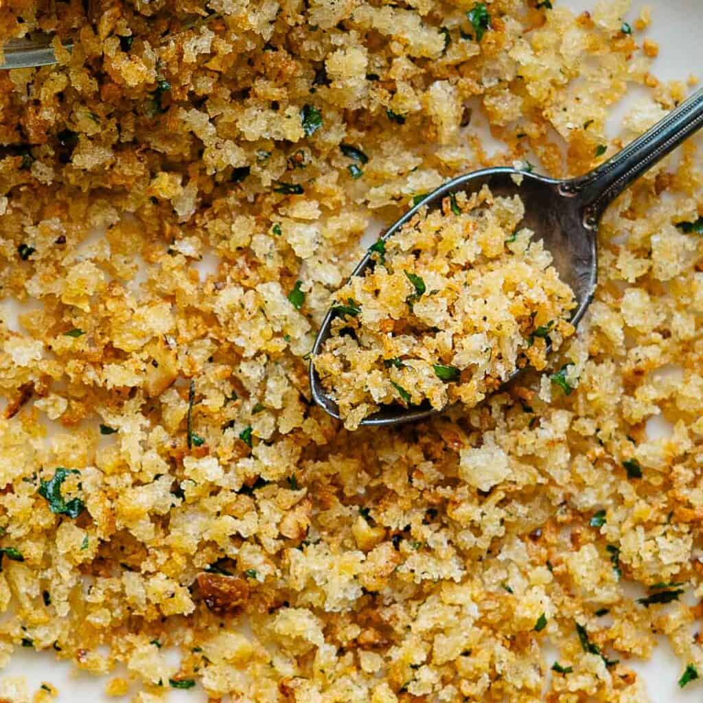 Close-up of a spoon scooping toasted pangrattato mixed with chopped herbs on a white surface. The golden brown and crispy breadcrumbs look delicious and aromatic.