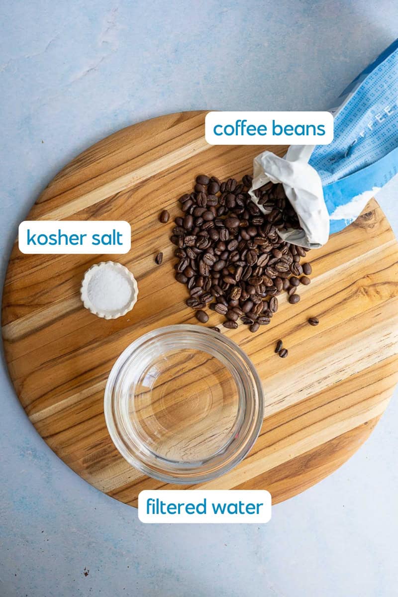 A wooden board displays a small bowl of kosher salt, a glass of filtered water, and a blue bag spilling coffee beans—key ingredients when learning how to make cold brew. Labels clearly identify kosher salt, filtered water, and coffee beans.