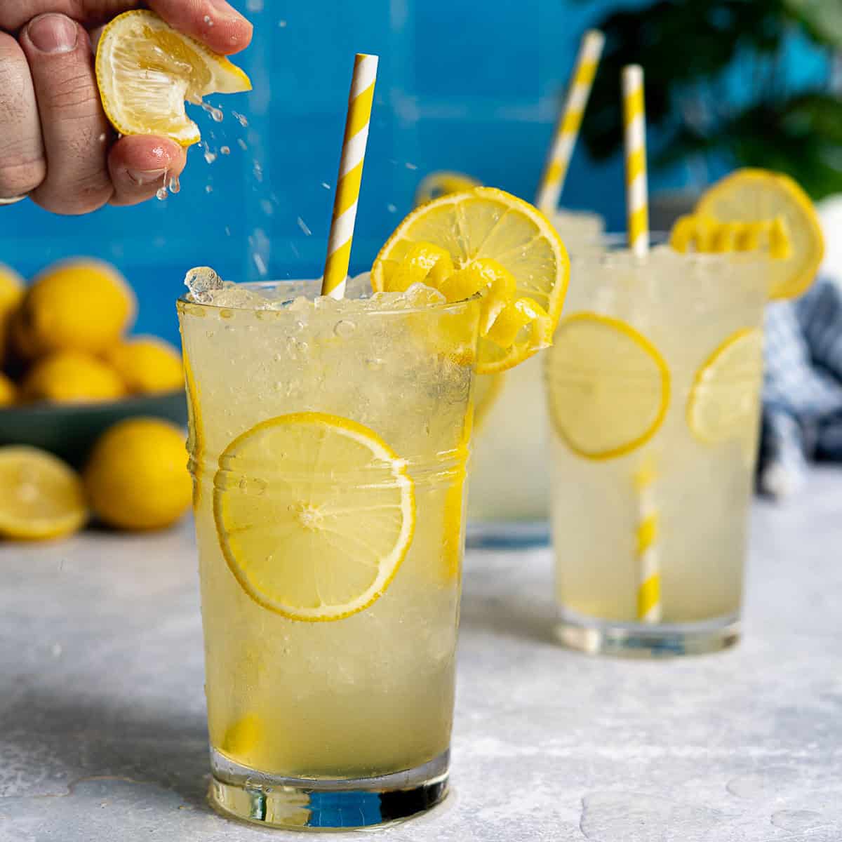 A hand squeezes a lemon wedge over a glass of Italian lemonade with ice, lemon slices, and a yellow-striped straw—capturing the refreshing spirit of Italian non alcoholic drinks. Two similar glasses and a bowl of lemons sit against a blue tiled wall.