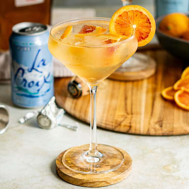 A blood orange spritz in a stemmed glass, garnished with a slice of orange, rests on a wooden coaster. Sliced oranges, a can of La Croix, and cocktail tools are blurred in the background.