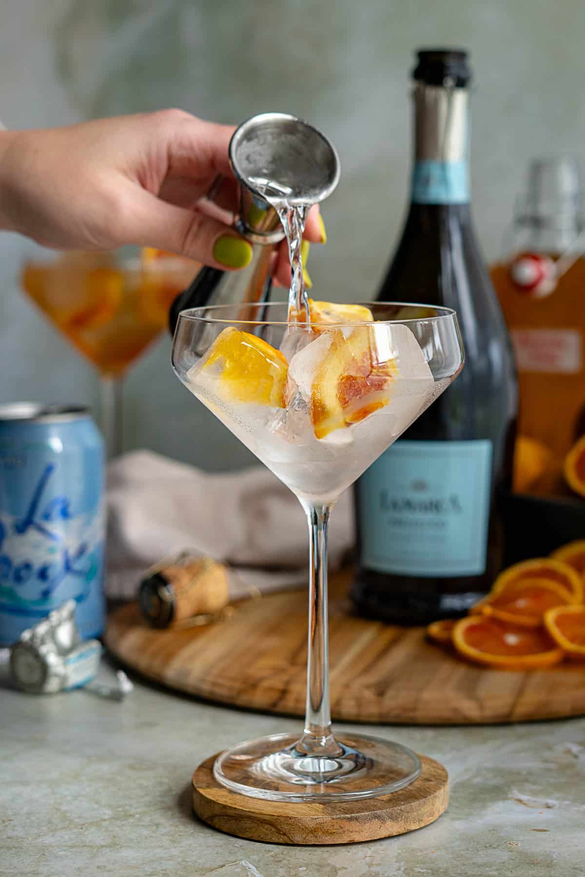 A hand pours soda water from a jigger into a cocktail glass with ice and orange slices, creating a refreshing blood orange spritz. In the background are a bottle of sparkling wine, La Croix, more orange slices, and a wooden surface.