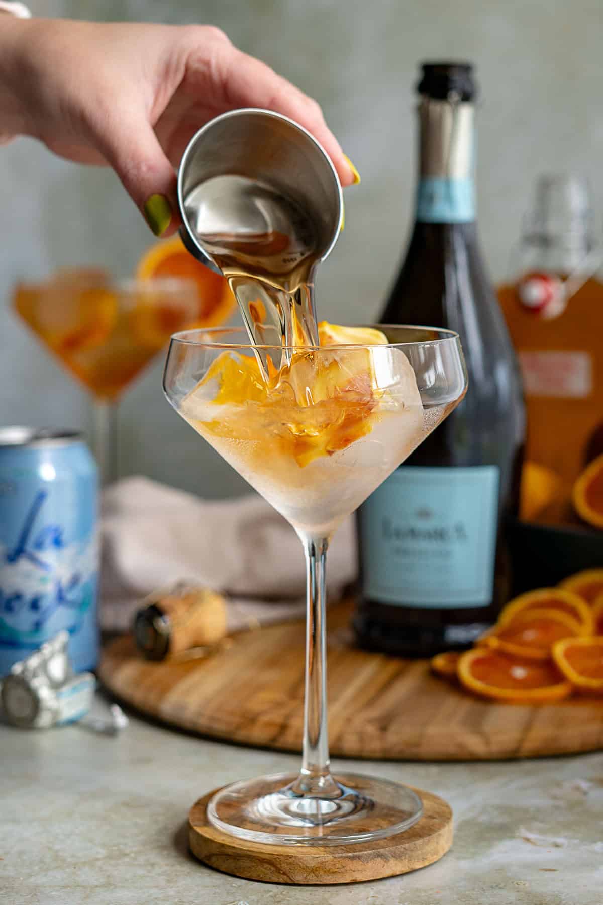 A hand pours blood orange liqueur from a metal jigger into a cocktail glass filled with ice and orange slices, crafting a refreshing blood orange spritz. A bottle of sparkling wine, a can of LaCroix, and orange slices sit on the table in the background.