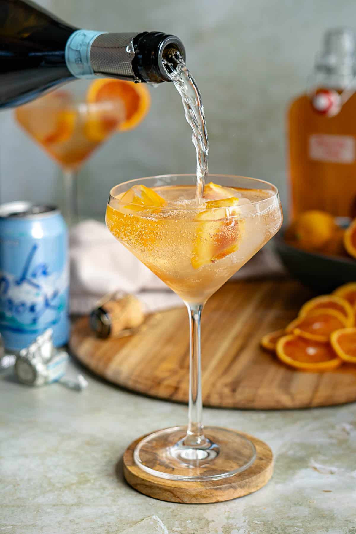 A blood orange spritz is being topped with prosecco in a glass filled with orange slices, set on a wooden coaster. In the background are an open can, a cork, a bottle, and sliced oranges on a cutting board.