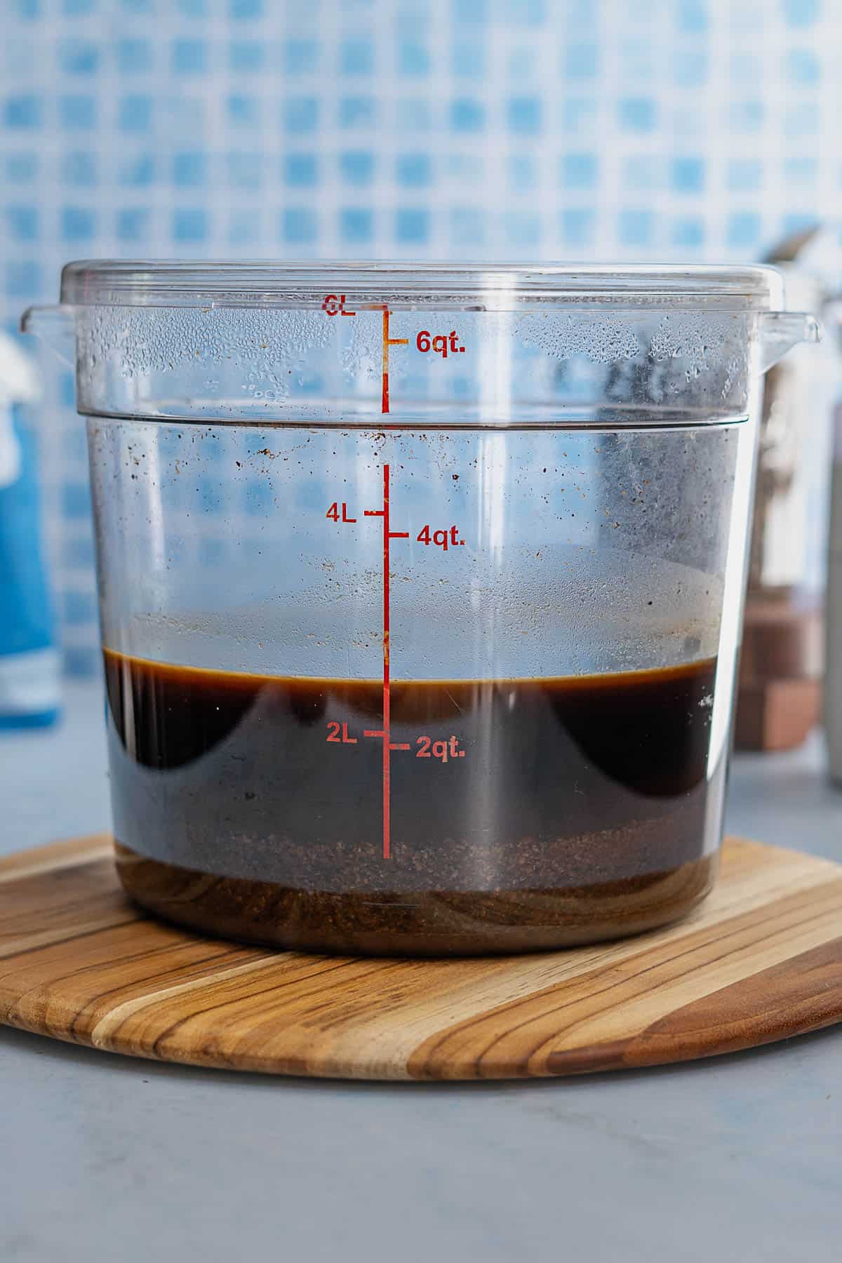 A clear plastic container with measurement markings holds a dark liquid with sediment at the bottom—like the first step in how to make cold brew—resting on a round wooden board against a blue tiled background.