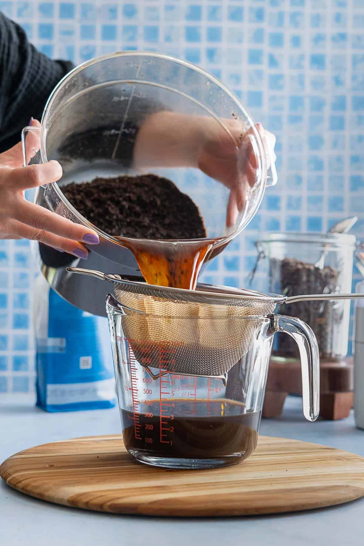 A person demonstrates how to make cold brew by pouring brewed coffee and grounds from a large bowl through a fine mesh strainer into a glass measuring cup on a wooden board, set against a blue-tiled background.