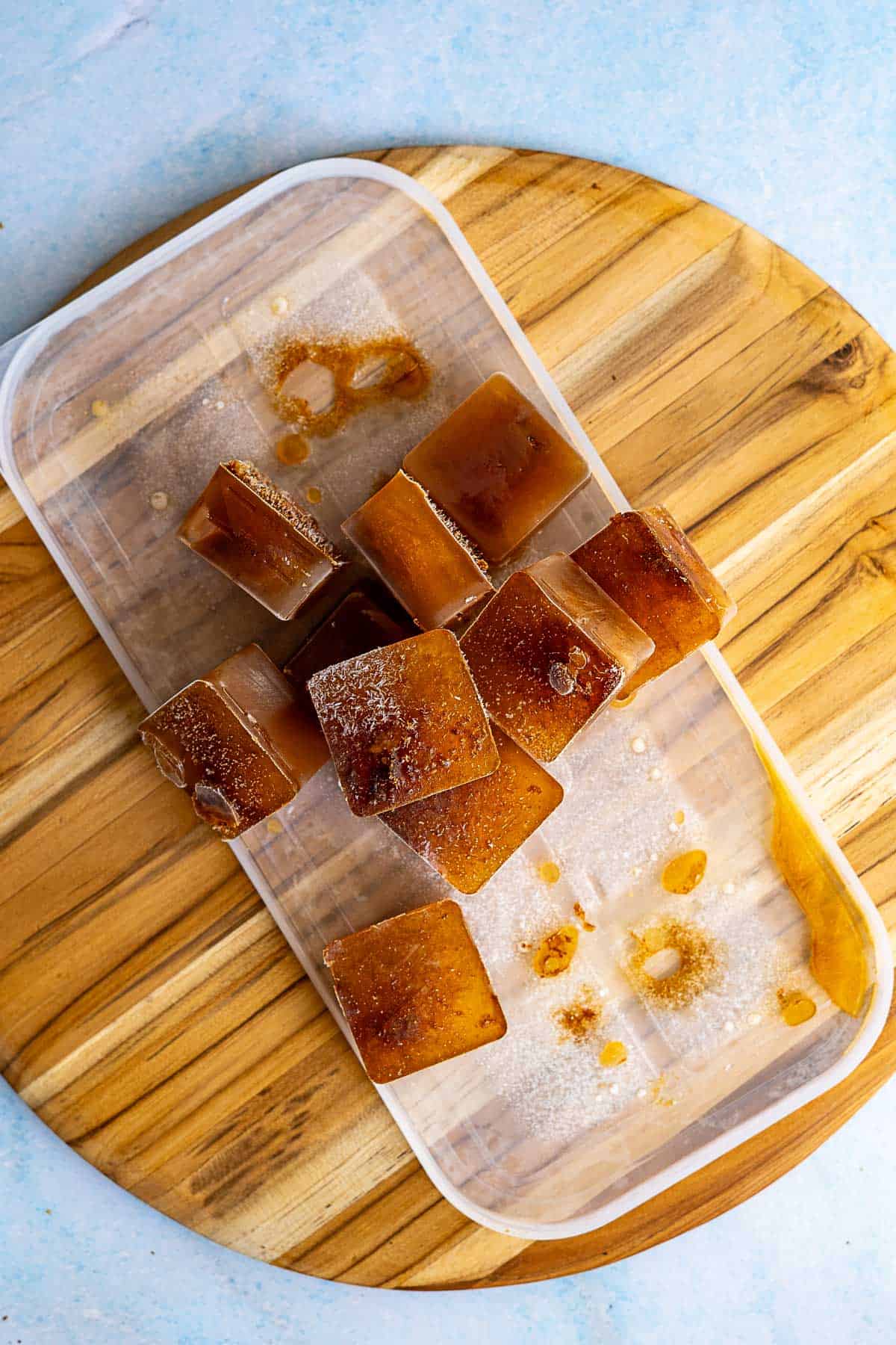 A plastic ice tray with several brown coffee ice cubes, some scattered on a round wooden board, demonstrates how to make cold brew easy and refreshing. The scene is set on a light blue background.