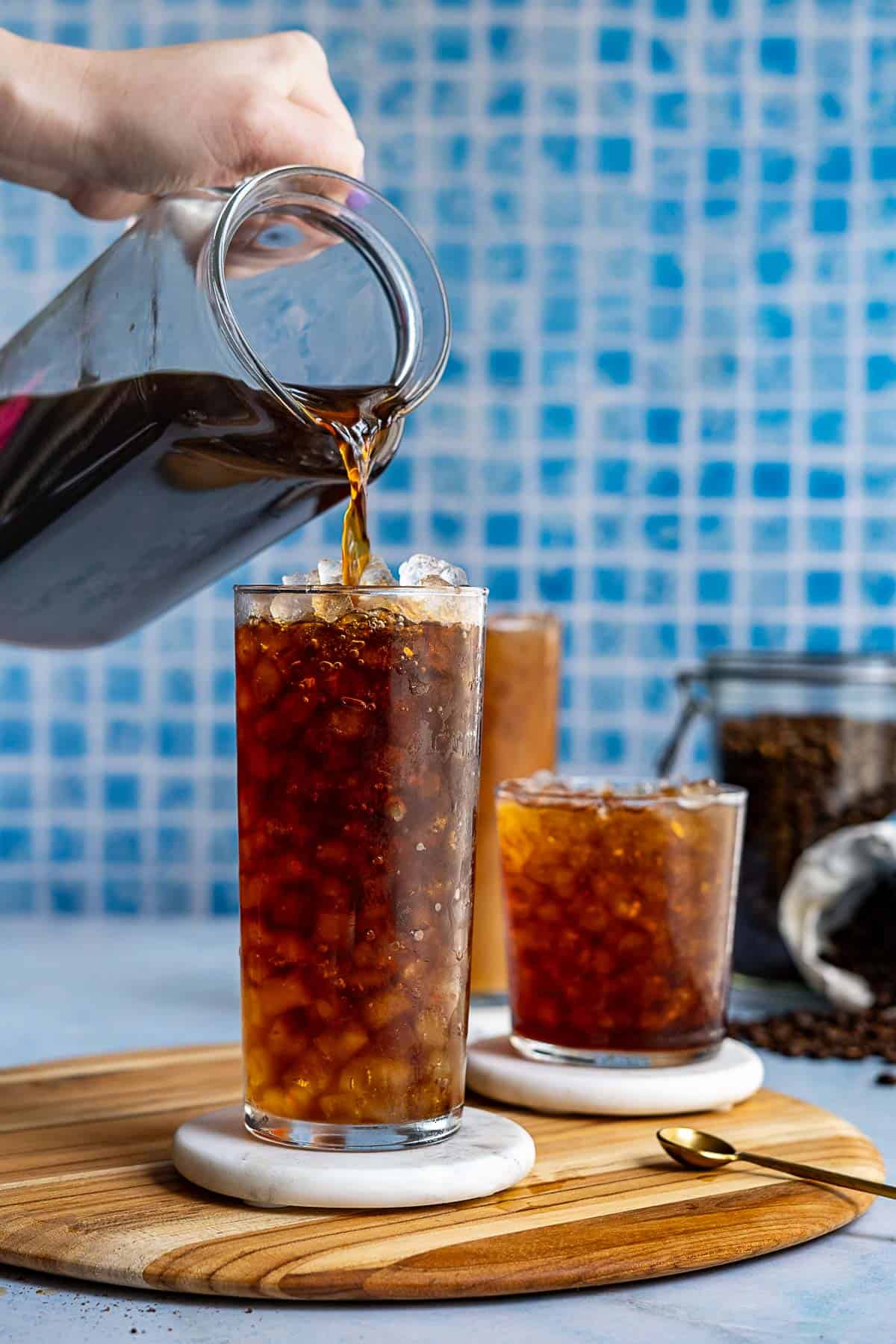 A hand pours iced coffee from a glass pitcher into a tall glass filled with ice, illustrating how to make cold brew, with two more glasses on a wooden board and a jar of coffee beans in the background.