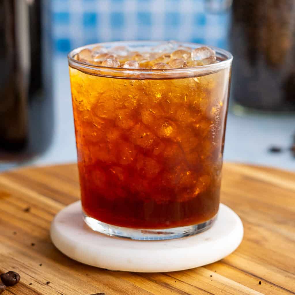 A glass filled with iced coffee sits on a white coaster atop a wooden surface, with crushed ice visible and a blurred blue background—perfect inspiration if you want to learn how to make cold brew at home.